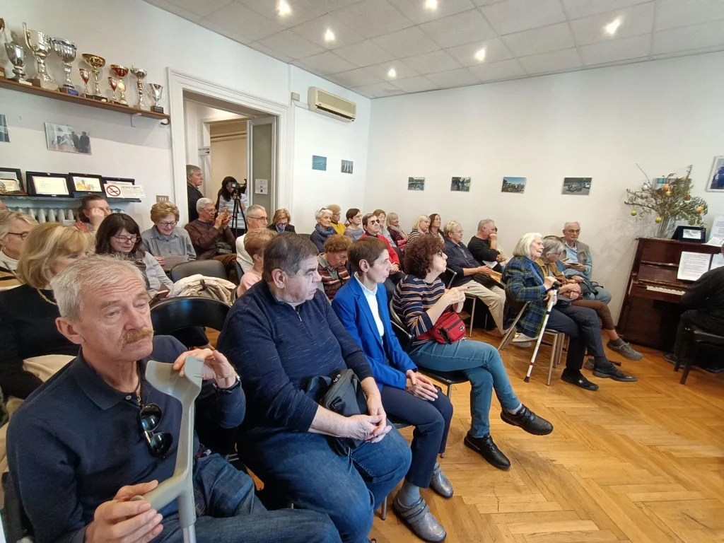 Un incontro del Circolo Tomè nella sala della sede: una trentina di persone sedute ascoltano, alcune con il bastone accanto. Alle pareti mensole con coppe e targhe; in fondo un pianoforte.
