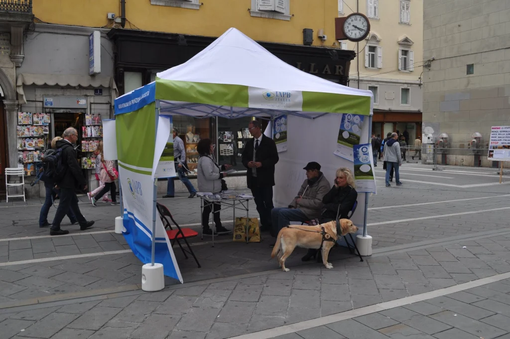 Giornata della prevenzione della cecità in una piazza di Trieste: sotto il gazebo bianco e verde di IAPB Italia i volontari parlano con i passanti. In primo piano un cane guida, un labrador chiaro, accanto a due persone sedute.