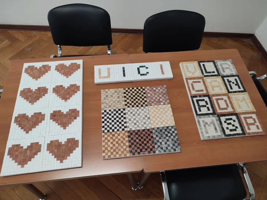 Mosaici realizzati dai soci, disposti su un tavolo: un pannello con otto cuori rossi su fondo bianco, uno a scacchiere di marmi colorati, uno con le lettere UICI e altri con lettere e numeri.